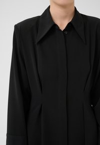 Camicia nera con bottoni, dotata di un ampio colletto, caratterizzata da una texture morbida e liscia e da pieghe frontali per un tocco di design in più.