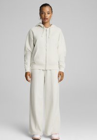 Sudadera de forro polar blanca con cierre y ajuste oversized, bolsillos tipo canguro y pantalones de pierna ancha a juego. Cuenta con una capucha y puños elásticos.