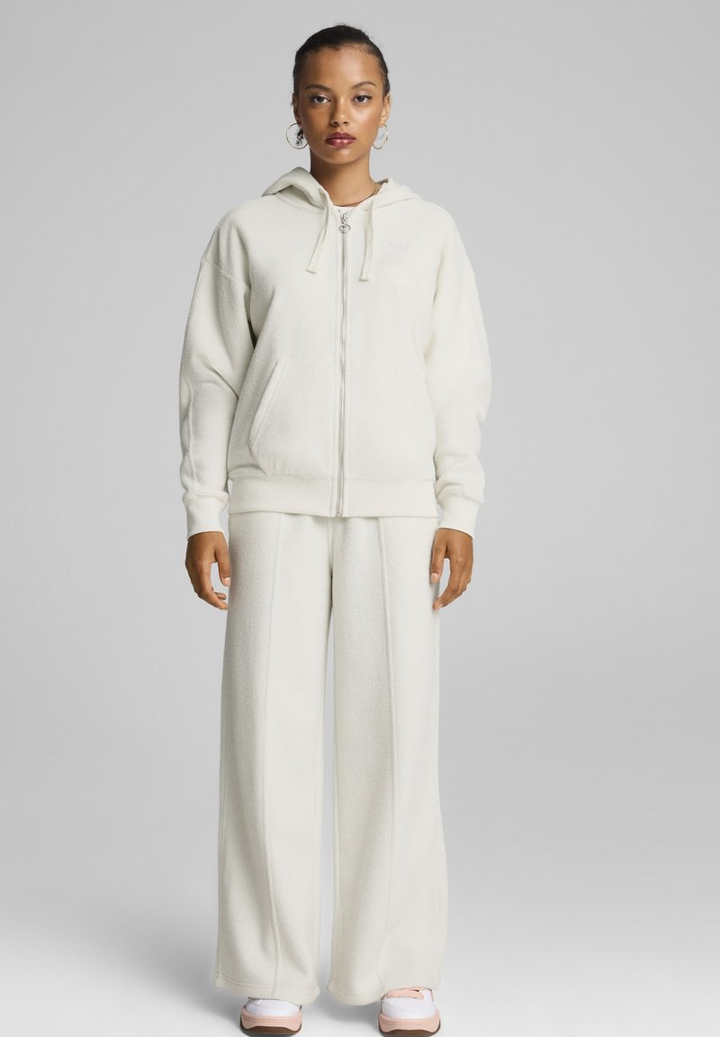 Sudadera de forro polar blanca con cierre y ajuste oversized, bolsillos tipo canguro y pantalones de pierna ancha a juego. Cuenta con una capucha y puños elásticos.