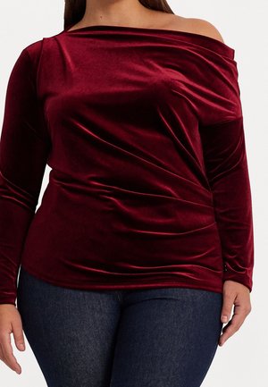 Frau trägt ein figurbetontes, langärmliges, schulterfreies Samtoberteil in tiefrot, kombiniert mit dunkelblauen Jeans.