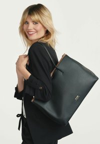 Czarna torba tote z teksturą i brązowymi akcentami, charakteryzująca się minimalistycznym designem i solidnym uchwytem, trzymana przez osobę w eleganckim stroju.