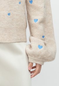 Beige wool sweater with ribbed cuffs, featuring scattered light blue heart patterns. Soft texture, cropped design. Fitted silhouette.