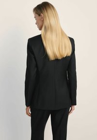 Czarna marynarka o dopasowanym kroju, z wyraźnie zaznaczonymi ramionami, eleganckim stylem i gładką fakturą. Posiada środkową szew i dopasowaną sylwetkę.