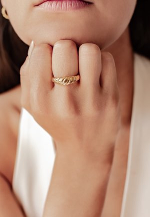 Goldring mit einem gedrehten Design, dargestellt auf einer Hand, die auf einem Kinn ruht. Die Hautstruktur ist sichtbar und der Hintergrund ist neutral.