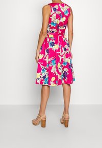 Blommig klänning i livfullt rosa med blå, gula och lila mönster. Ärmlös design med en figursydd midja och utsvängd kjol. Sandaler med träklack.