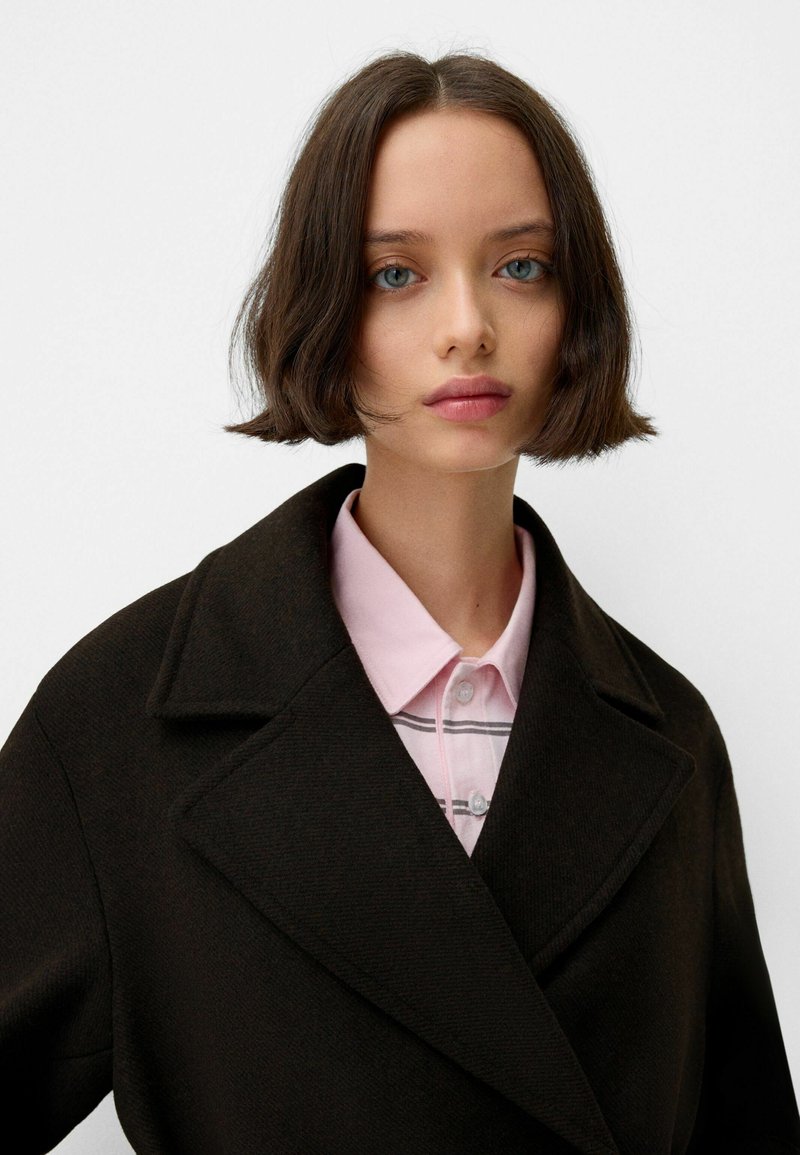 Czarny wełniany płaszcz z wykładanym kołnierzem na jasnoróżowej koszuli z kołnierzykiem, w poziome paski. Gładka tekstura i dopasowany krój.