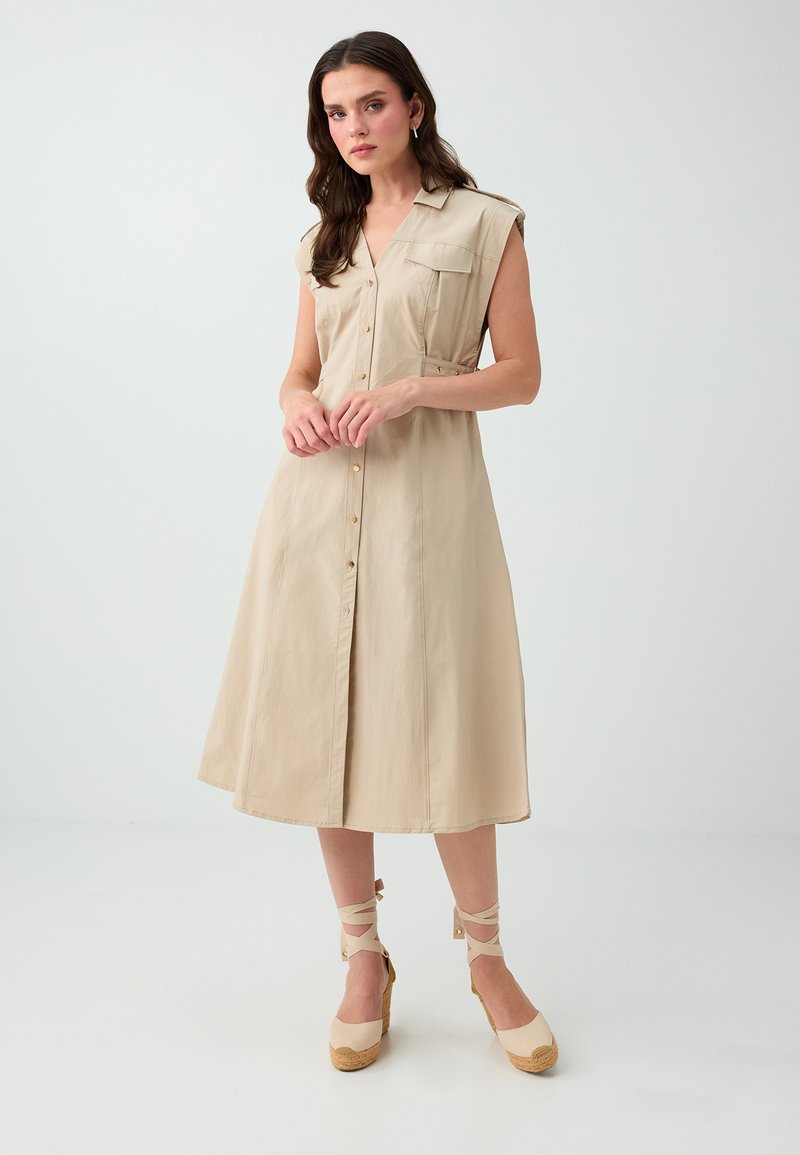 Femme aux longs cheveux foncés portant une robe midi beige sans manches boutonnée et des chaussures compensées beige avec des lacets aux chevilles, debout sur un sol blanc.