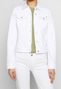 Nainen, jolla on päällä valkoinen farkkutakki hopeisilla nappeilla, vaaleanvihreä toppi ja valkoiset housut, yksivärisen taustan edessä.