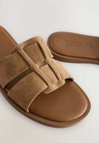 Smeđe suede natikače s dva široka remena, s geometrijskim izrezanim dizajnom i glatkom smešnom podlogom. Ravna potplat, ležeran stil.