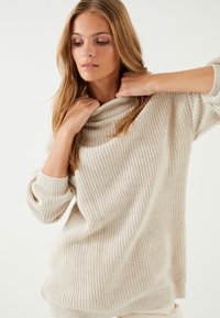 Beżowy sweter wykonany na drutach z luźnym dekoltem typu cowl, długimi rękawami i prążkowaną fakturą. Posiada luźny krój i subtelne poziome paski.