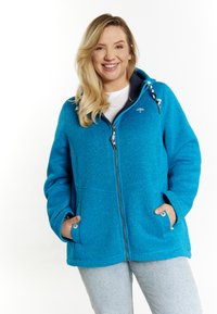 Giacca in pile blu con zip e cappuccio, dotata di tasche laterali, lacci e finitura testurizzata. Indossata con una t-shirt bianca sotto.