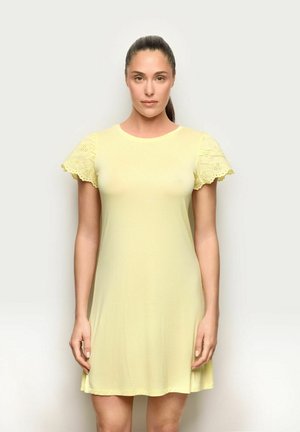 Femme aux cheveux foncés en queue de cheval, portant une courte robe jaune avec des manches courtes en dentelle ajourée, debout devant un fond clair uni.