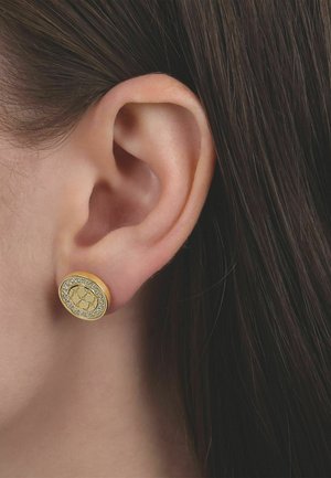 Nahaufnahme eines Ohrs mit kurzen dunklen Haaren, das einen runden goldenen Ohrstecker trägt, der ein zentrales Muster zeigt und von kleinen klaren Steinen umgeben ist.