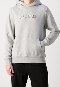 Felpa grigia con tasca frontale, con la scritta "HILFIGER NEW YORK" in blu navy e rosso sul petto. Tessuto spesso e polsini a coste.