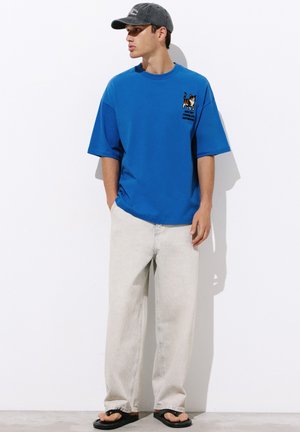 Mann mit blauem T-Shirt mit Cartoon-Katze, heller Jeans, schwarzen Sandalen und grauer Kappe steht vor einer weißen Wand.
