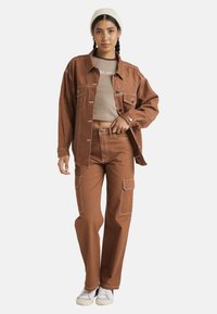 Giacca di jeans marrone con tasche frontali e cuciture bianche, abbinata a un top corto marrone chiaro e pantaloni cargo larghi coordinati. Sneakers bianche.