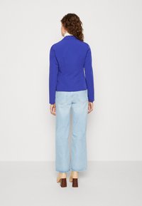 Modrý přiléhavý blejzr se strukturovaným střihem, doplněný světle modrými širokými džínami a béžovými botami na blokovém podpatku. Jednoduché pozadí.