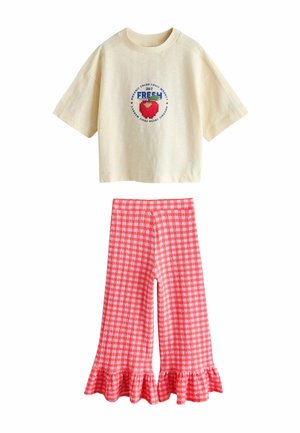 Kremowy krótki crop top z krótkim rękawem, ozdobiony pikselową czerwoną truskawką i napisem, zestawiony z szerokimi spodniami w czerwono-białą kratę zakończonymi falbankami.