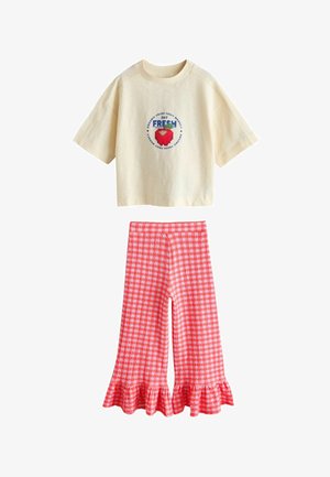 Krém színű, rövid ujjú, haspóló, melyen pixeles vörös eper és felirat látható, piros-fehér kockás, bőszárú nadrággal, fodros szegélyekkel kombinálva.