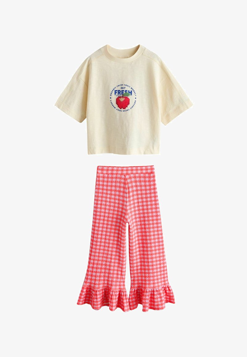 Krémové krátké tričko s krátkým rukávem a pixelovým červeným jahodovým motivem a textem, doplněné červenobílými širokými kalhotami s rozparkem a volánky na lemu.