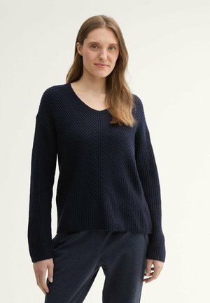 Granatowy sweter z dekoltem w serek, prążkowaną teksturą i detalami w wzór zygzaka. Długie rękawy i luźny krój podkreślają jego swobodny styl.