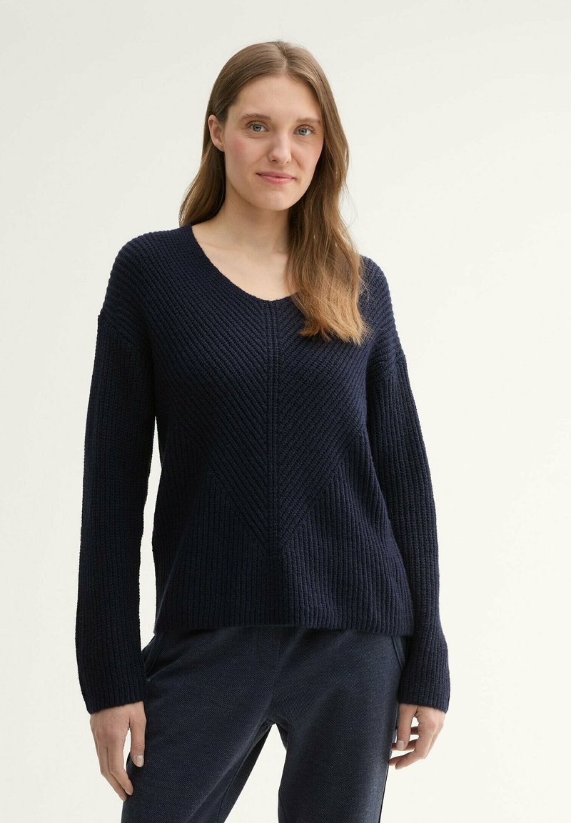 Granatowy sweter z dekoltem w serek, prążkowaną teksturą i detalami w wzór zygzaka. Długie rękawy i luźny krój podkreślają jego swobodny styl.
