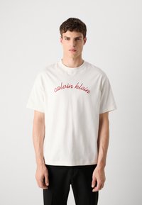 White cotton t-shirt with short sleeves, featuring red "calvin klein" text in cursive across the chest. Simple, relaxed fit design.