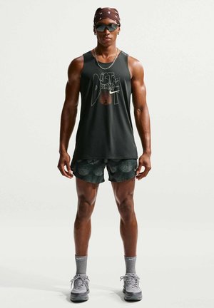 Atletyczna osoba nosząca czarną bezrękawnikową koszulkę Nike, wzorzyste szorty, szare skarpetki Nike, trampki, okulary przeciwsłoneczne i brązową bandanę, stojąca prosto.