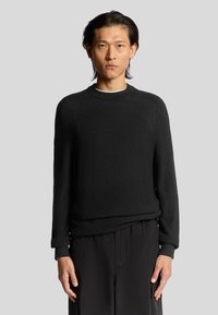 Maglione nero in maglia con polsini e scollatura a coste, caratterizzato da una vestibilità comoda e una texture liscia. Indossato sopra pantaloni scuri.