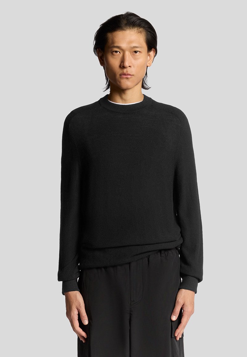 Maglione nero in maglia con polsini e scollatura a coste, caratterizzato da una vestibilità comoda e una texture liscia. Indossato sopra pantaloni scuri.