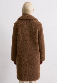 Cappotto marrone e peloso con colletto alto e maniche lunghe. Il tessuto presenta una superficie texture e morbida. L'orlo raggiunge la metà della coscia.