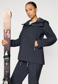 Geacă de schi din țesătură bleumarin cu finisaj impermeabil, având glugă, buzunare cu fermoar și un design elegant. Schiuri ținute în mâna stângă.
