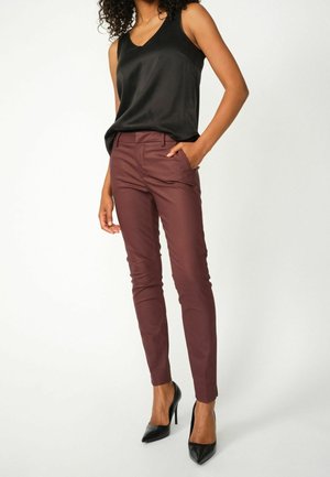 Kvinde iført en sort ærmeløs top, bordeaux slim-fit bukser og sorte højhælede sko, stående med den ene hånd i lommen.