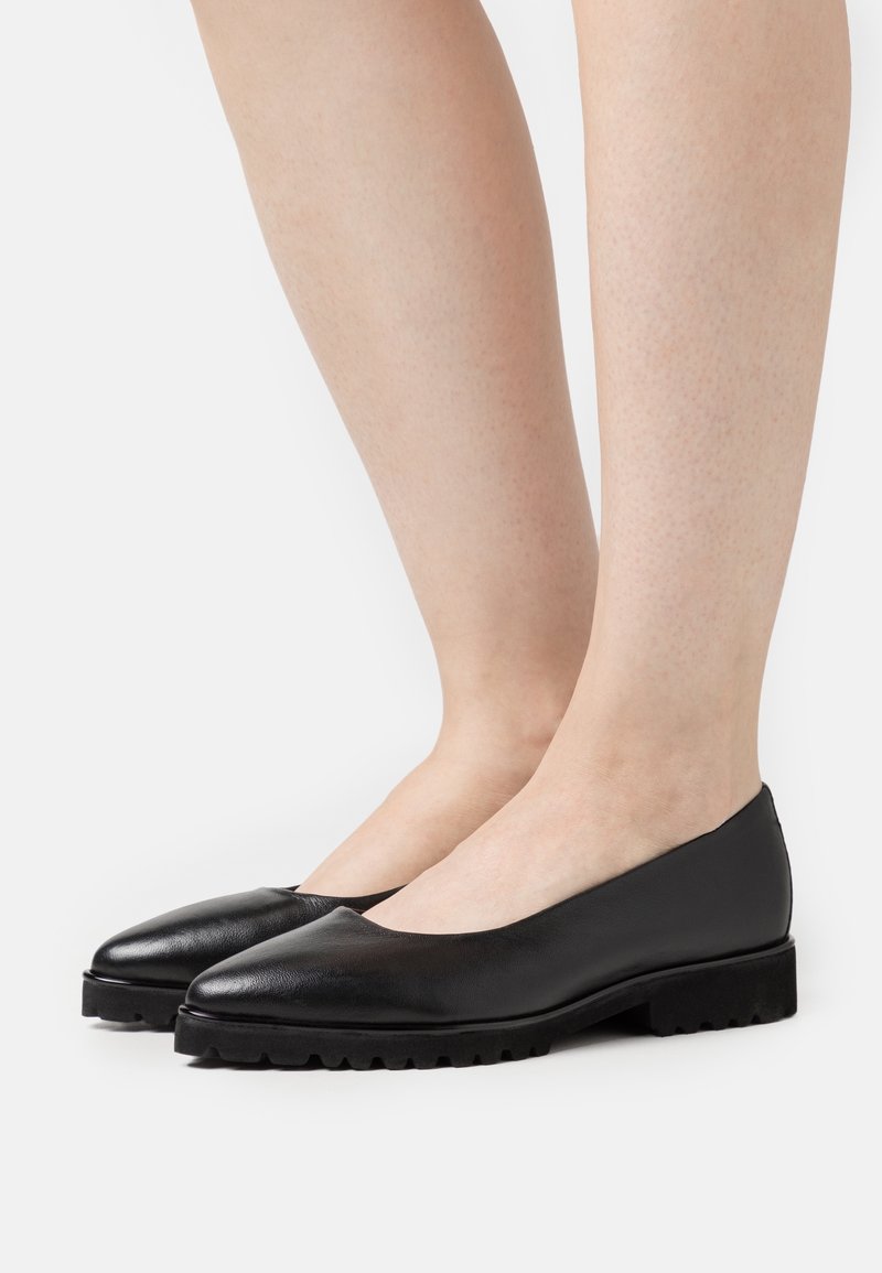 Jambes portant des ballerines en cuir noir à bouts arrondis et semelles épaisses à crampons sur fond blanc.