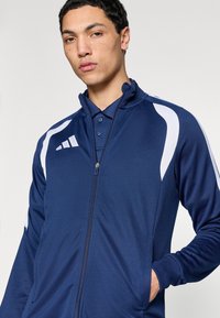 Μπλε ναυτικό μπουφάν με φερμουάρ και λευκές λεπτομέρειες, κατασκευασμένο από ομαλό ύφασμα. Διαθέτει ανασηκωμένο γιακά και πλαϊνές τσέπες. Το λογότυπο της Adidas είναι ορατό στο στήθος.
