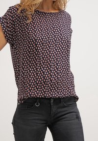 Mörkblå blus med korta ärmar, med ett geometriskt mönster i ljus persika. Lättviktsmaterial, avslappnad passform, rundad halsringning. Svarta jeans synliga.