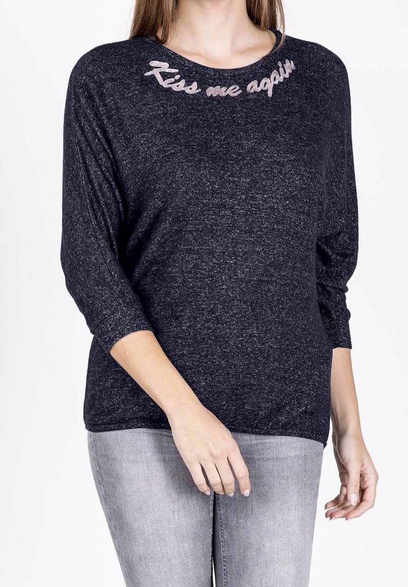 Femme portant un pull gris foncé chiné avec le texte rose "Embrasse-moi encore" brodé près de l'encolure, associé à un jean gris clair.