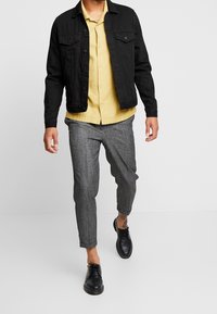 Černá džínová bunda, žlutá košile na knoflíky, šedé vzorované zúžené kalhoty a černé boty. Neformální outfit s vrstvenými texturami.
