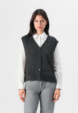 Young woman stands confidently, wearing a sleeveless dark gray vest over a white collared shirt and gray jeans, with a neutral background.