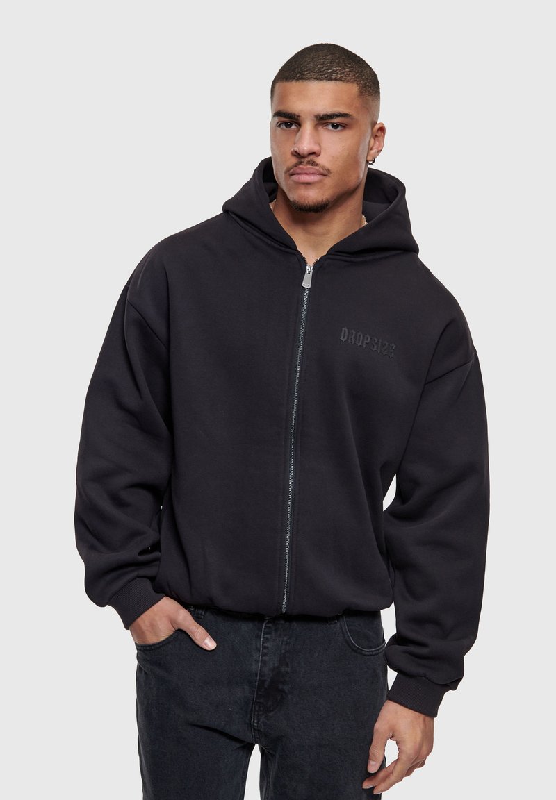 Hombre con sudadera negra con cremallera y texto "Dropsix" en el pecho, con las manos en los bolsillos, de pie frente a un fondo liso gris claro.