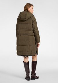 Oliwkowo-zielona pikowana kurtka z kapturem, z przeszyciami w kwadraty i bocznym rozcięciem. Nosi się ją na wzorzystej tkaninie, w zestawieniu z wysokimi, błyszczącymi kozakami do kolan.