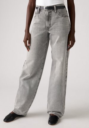 Personne portant un jean gris délavé taille haute à jambes larges, une ceinture noire, un haut blanc et des chaussures plates noires sur un fond uni.