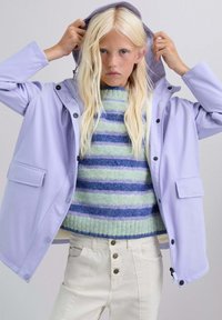 Chaqueta impermeable de color lavanda claro con capucha, cierre con botones de presión y bolsillos delanteros. Lleva puesta sobre un suéter borroso a rayas y pantalones blancos.