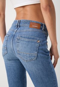 Gros plan d'une personne portant un jean denim Mos Mosh bleu clair avec des poches arrière et un top court blanc, montrant le bas du dos et la taille.
