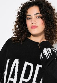 Schwarzer Pullover mit großem weißen "HAPPY"-Aufdruck. Gerippter Rundhalsausschnitt und Säume. Strukturierter Stoff mit einem weichen, flauschigen Erscheinungsbild. Weiße Tropfenakzente an den Ärmeln.