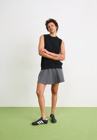 Svart ermeløs topp med teksturert design, parret med en grå plisserte skjørt og svarte joggesko med hvite striper. Grønn gulvbakgrunn.