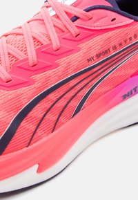 Κοντινό πλάνο ενός έντονα ροζ αθλητικού παπουτσιού με υφή υφάσματος, μαύρο σχέδιο «swoosh» και το κείμενο «MY SPORT IS HYRO» στο πλάι.