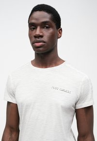 T-shirt de algodão cinza claro com decote redondo, apresentando "JUST CAVALLI" bordado a preto na área do peito esquerdo.