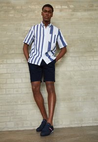 Kortärmad randig skjorta i blått och vitt, kombinerad med marinblå shorts. Har en knäppt ficka och mörka sneakers med röd accent.