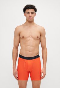 Oransje boksershorts med en svart elastisk midjebånd som har ordet "UNDER" i fete bokstaver, laget av mykt, elastisk materiale.